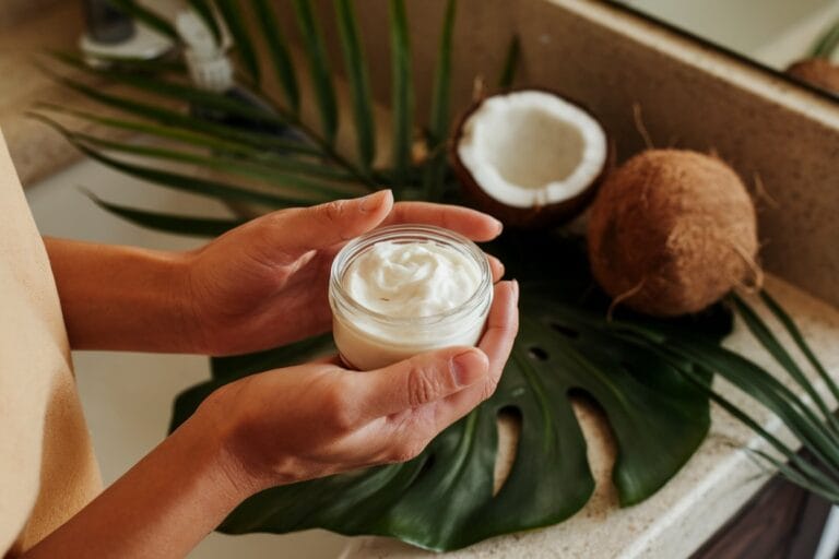 Close-Up Lifestyle Photograph Of A Woman'S Hands Holding A Small Glass Jar Filled With Creamy White Homemade Hair Conditioner, Soft Natural Lighting, Bathroom Counter With Tropical Leaves And Coconut In Background, Warm And Inviting Atmosphere, Editorial Style Photography