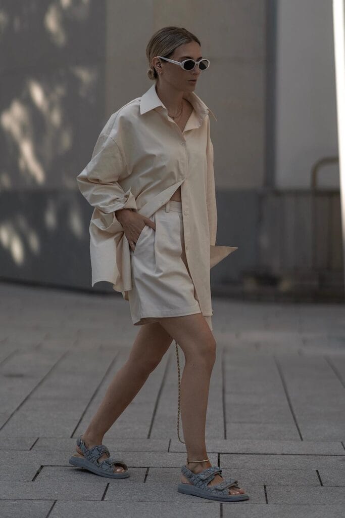 How To Style Oversized Shirts Like A Fashion Blogger 15 Woman In Oversized Beige Shirt And Matching Shorts, Walking On A City Street, Wearing Sunglasses And Sandals.