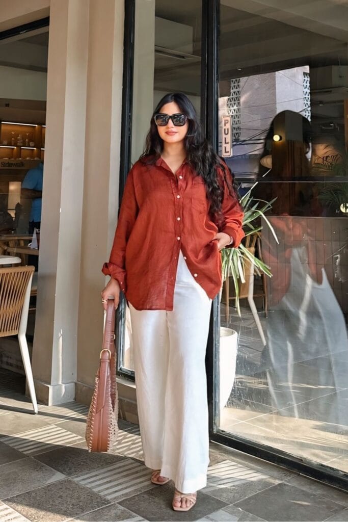 How To Style Oversized Shirts Like A Fashion Blogger 19 A Woman Standing In Front Of A Café, Wearing A Rust-Colored Oversized Button-Up Shirt And White Wide-Leg Pants, Carrying A Stylish Handbag And Wearing Sunglasses.