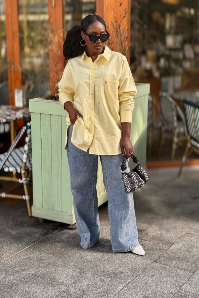 How To Style Oversized Shirts Like A Fashion Blogger 9 Woman Wearing A Yellow Oversized Shirt And Wide-Leg Jeans, Holding A Small Handbag, Posing Casually Outdoors.