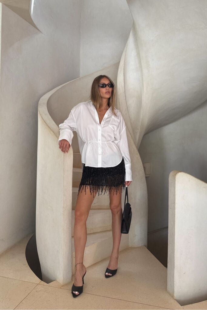 How To Style Oversized Shirts Like A Fashion Blogger 10 Woman Wearing An Oversized White Shirt With A Tied Waist, Black Fringe Mini Skirt, And Sunglasses, Standing On A Spiral Staircase In A Stylish Setting.
