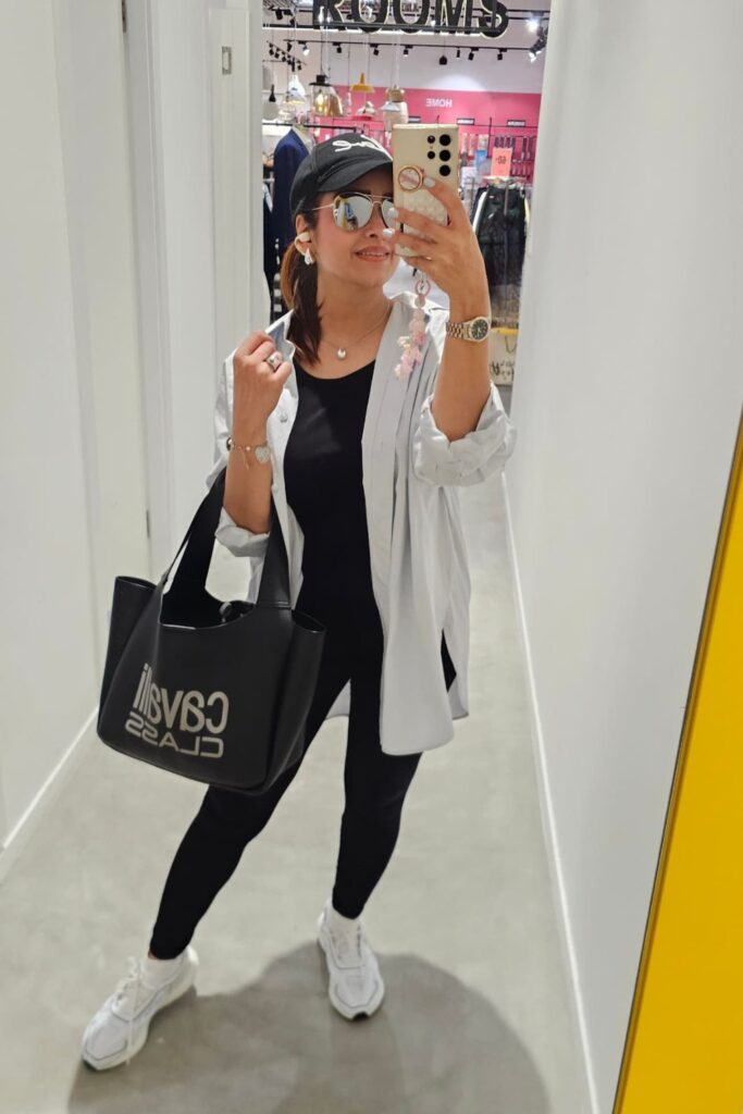 How To Style Oversized Shirts Like A Fashion Blogger 13 A Person Taking A Mirror Selfie Wearing An Oversized White Shirt, Black Top, And Black Leggings, Accessorized With A Black Cap, Sunglasses, And A Black Bag With A Logo.