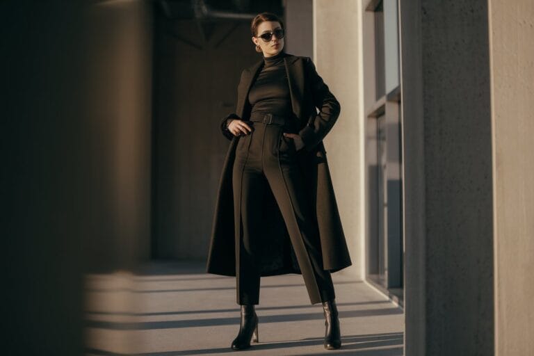 A Confident Person Of Average Height Standing In An Urban Winter Setting, Wearing A Monochrome Black Outfit Consisting Of High-Waisted Tailored Pants, Fitted Black Turtleneck, Long Wool Coat Hitting Below The Knee, And Heeled Chelsea Boots. The Styling Makes Them Appear Taller And Elegant. Natural Window Lighting From The Side, Modern Minimalist Interior With Concrete Walls, Full-Body Shot Showing Complete Proportions, Fashion Editorial Style, Sharp Focus, Professional Photography, Clean Composition Emphasizing Vertical Lines And Sleek Silhouette. Warm Color Grading With Cool Winter Tones.